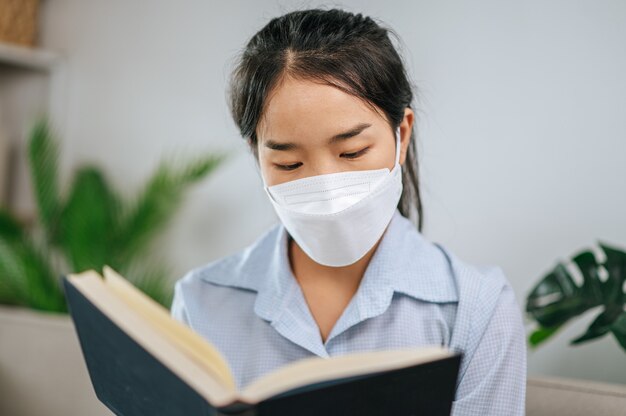 Jonge aziatische vrouw met gezichtsmasker zittend op de bank in de woonkamer, ze leest boek tijdens quarantaine covid-19 zelfisolatie thuis