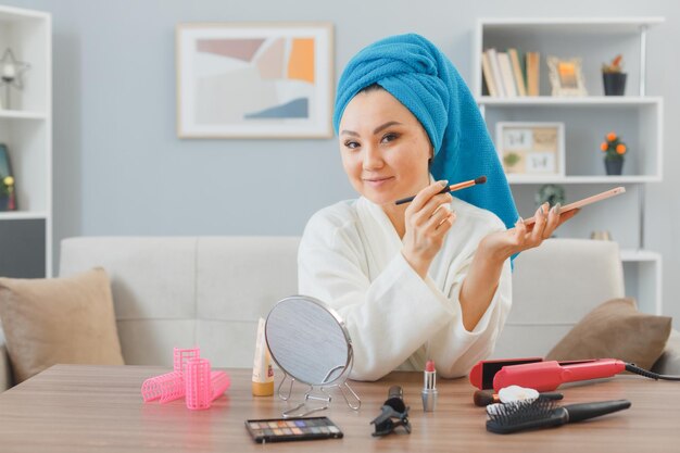 jonge aziatische vrouw met handdoek op haar hoofd zittend aan de kaptafel thuis interieur oogschaduw toe te passen met palet glimlachen doen ochtend make-up routine schoonheid en gezichtscosmetica concept