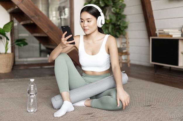 Jonge Aziatische vrouw met waterfles luisteren muziek of podcast in koptelefoon tijdens fitness thuis...