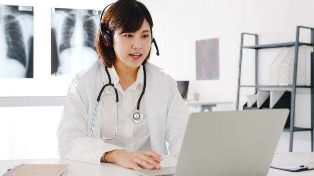 Jonge Aziatische vrouwelijke arts in wit medisch uniform met stethoscoop met behulp van computerlaptop pratende videoconferentiegesprek met patiënt aan balie in gezondheidskliniek of ziekenhuis.