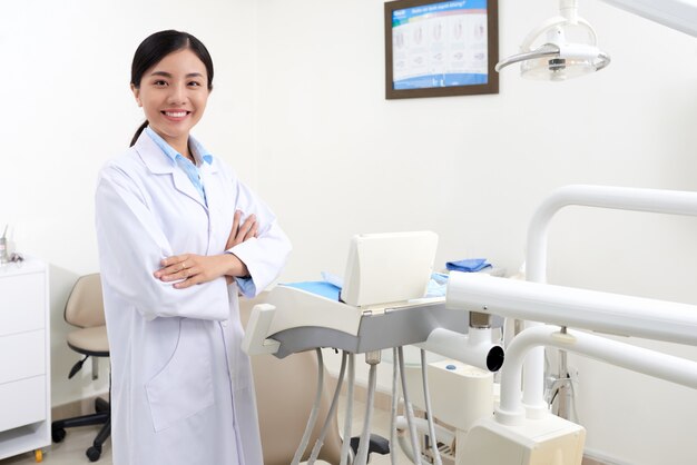 Jonge Aziatische vrouwelijke tandarts in witte jas poseren in kliniek naast apparatuur