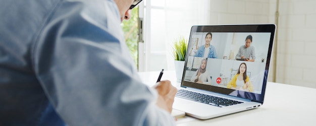 Jonge aziatische zakenman die laptop met behulp van praat met collega's over plan in videogesprekvergadering terwijl het werken vanuit huis in de woonkamer. zelfisolatie, sociale afstand nemen, quarantaine voor coronaviruspreventie.