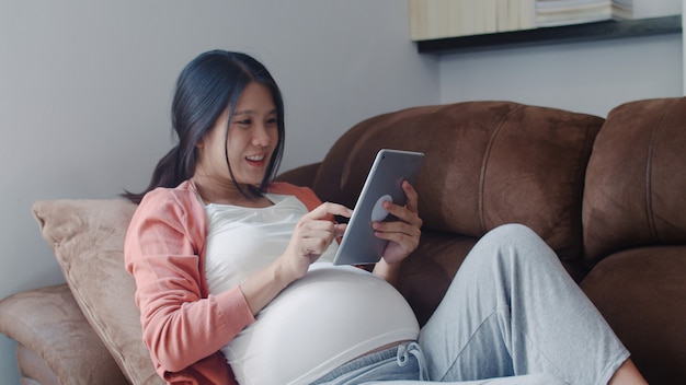 Jonge aziatische zwangere vrouw die de zwangerschapsinformatie van het tabletonderzoek gebruikt. mamma die het gelukkige positief en vreedzame glimlachen voelen terwijl zorg haar kind thuis liggend op bank in woonkamer.