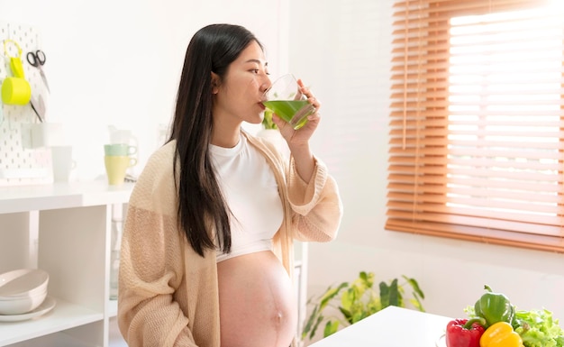 Jonge Aziatische zwangere vrouw die groen groentesap drinkt in de keuken en eet om een goede gezondheid te behouden tijdens de zwangerschap tot de bevalling