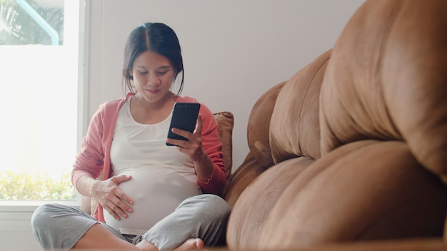 Jonge Aziatische zwangere vrouw met behulp van mobiele telefoon zoeken zwangerschap informatie. Mamma die het gelukkige positief en vreedzame glimlachen voelen terwijl zorg haar kind thuis liggend op bank in woonkamer.