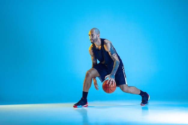 Jonge basketbalspeler van team dragen sportkleding training, oefenen in actie, beweging geïsoleerd op blauwe muur in neonlicht. Concept van sport, beweging, energie en dynamische, gezonde levensstijl.