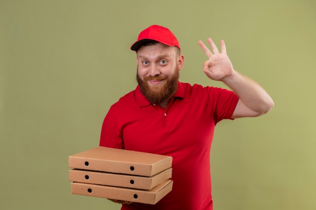 Jonge, bebaarde bezorger in rood uniform en pet bedrijf stapel pizzadozen lachend met blij gezicht weergegeven: ok teken