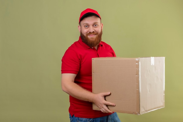 Jonge, bebaarde bezorger in rood uniform en pet met kartonnen doos camera kijken met zelfverzekerde glimlach