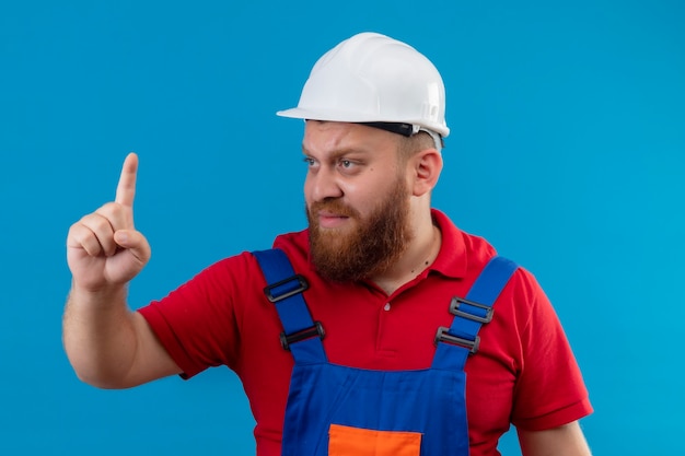 Gratis foto jonge, bebaarde bouwersmens in bouwuniform en veiligheidshelm op zoek opzij verward wijzende vinger omhoog