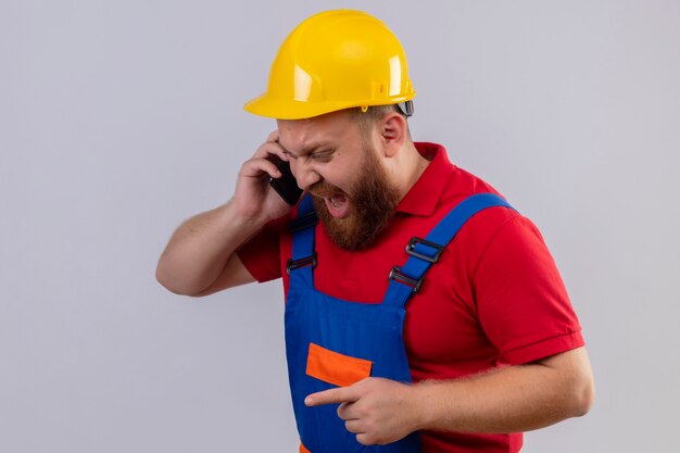 Jonge, bebaarde bouwersmens in bouwuniform en veiligheidshelm schreeuwen tijdens het gesprek op mobiele telefoon met agressieve uitdrukking