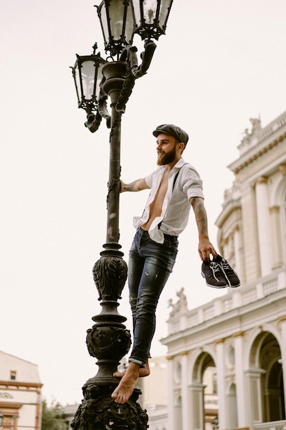 Jonge, bebaarde getatoeëerde man. Een romantische man in een wit overhemd, pet en bretels loopt door de stad. draait om een lantaarnpaal. Peaky Blinders. ouderwets, retro.