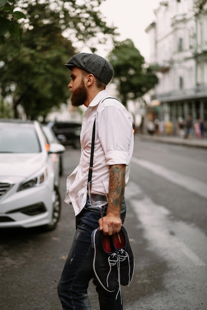 Jonge, bebaarde getatoeëerde man. Een romantische man in een wit overhemd, pet en bretels loopt door de stad. draait om een lantaarnpaal. Peaky Blinders. ouderwets, retro.