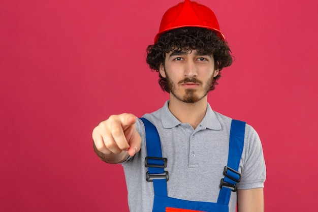 Jonge, bebaarde knappe bouwer bouw uniform dragen en veiligheidshelm verwijtend kijken naar de camera en een vinger op je wijzen over geïsoleerde roze muur