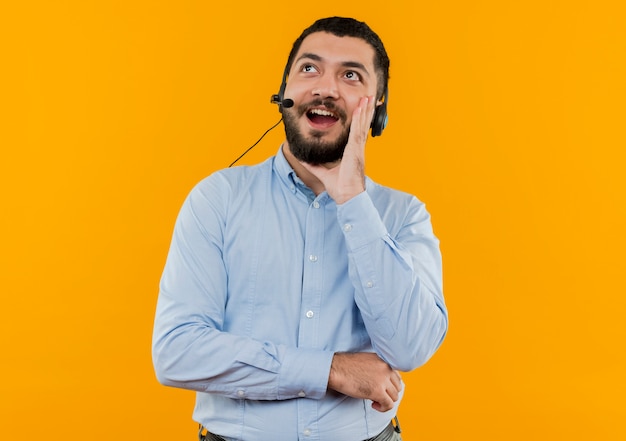 Jonge, bebaarde man in blauw shirt met koptelefoon met microfoon iemand bellen met de hand in de buurt van mond glimlachen