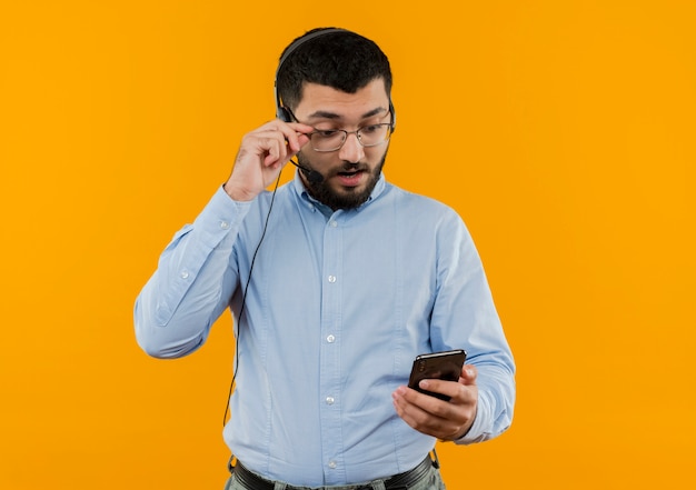 Jonge, bebaarde man in blauw shirt met koptelefoon met microfoon kijken naar zijn smartphonescherm zijn bril afzetten