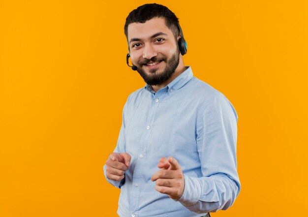 Jonge, bebaarde man in blauw shirt met koptelefoon met microfoon pointign met wijsvingers op camera vrolijk lachend