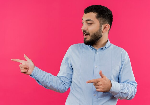 Jonge, bebaarde man in blauw shirt wijzend met wijsvinger naar de zijkant glimlachend gelukkig en positief