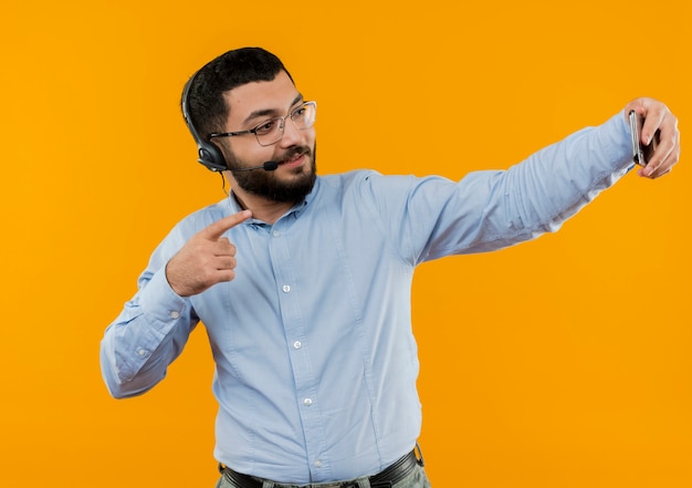 Jonge, bebaarde man in glazen en blauw shirt met koptelefoon met microfoon doet selfie met zijn smartphone glimlachen