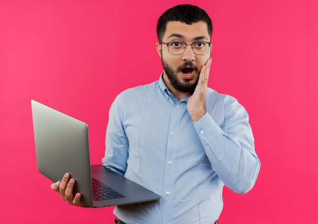 Jonge, bebaarde man in glazen en blauw shirt met laptop wordt verrast en verbaasd