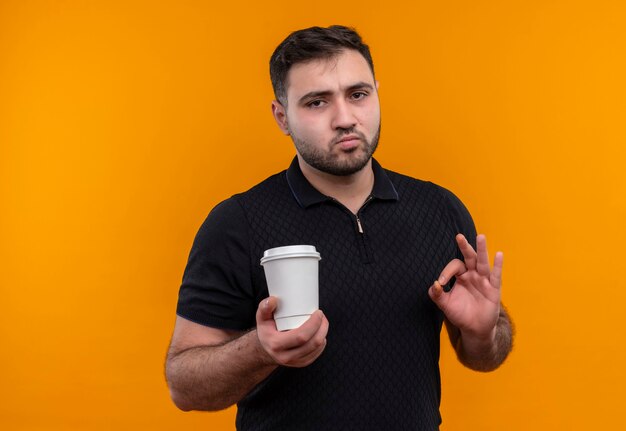 Jonge, bebaarde man in zwart shirt met koffiekopje ok teken op zoek zelfverzekerd te doen