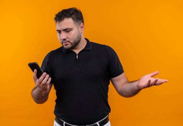 Jonge, bebaarde man in zwart shirt met smartphone op zoek verward en erg angstig