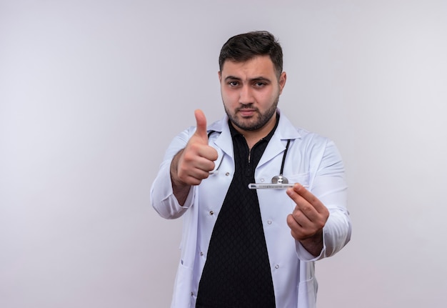 Jonge, bebaarde mannelijke arts die witte jas met de thermometer van de stethoscoopholding draagt ?? die camera bekijkt die duimen met gelukkig gezicht toont