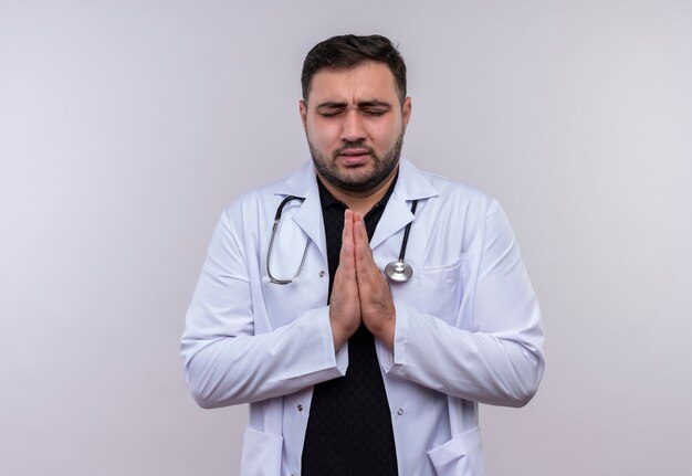 Jonge, bebaarde mannelijke arts die witte jas met een stethoscoop draagt die de wapens bij elkaar houdt, bidt en bedelt met hoop uitdrukking