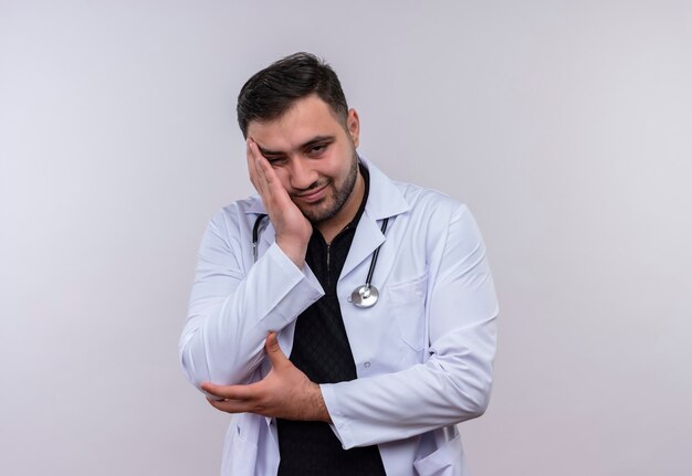 Jonge, bebaarde mannelijke arts die witte jas met een stethoscoop draagt die er moe en verveeld uitziet, zijn hoofd op de arm leunend