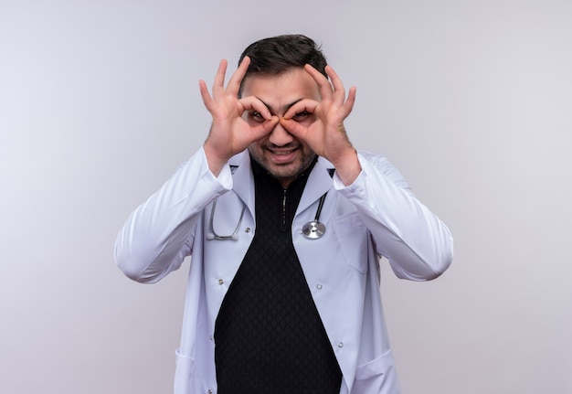 Jonge, bebaarde mannelijke arts die witte jas met een stethoscoop draagt die ok tekens met vingers doet als verrekijker die door vingers kijkt die pret heeft glimlachen