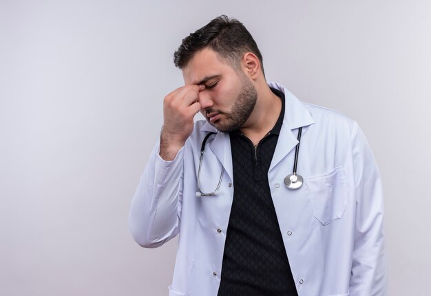 Jonge, bebaarde mannelijke arts die witte jas met een stethoscoop draagt die op zoek moe en verveeld neus tussen gesloten ogen aanraakt