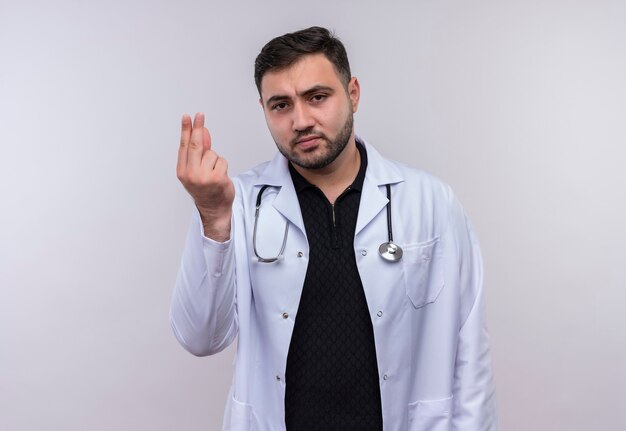 Jonge, bebaarde mannelijke arts die witte jas met stethoscoop draagt die camera bekijkt die het gebaar van het vingersgeld, vraagt om geld