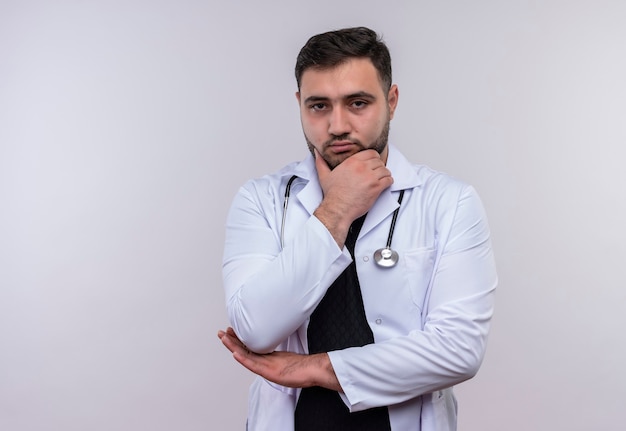 Jonge, bebaarde mannelijke arts die witte jas met stethoscoop draagt die camera met hand op kin met peinzende uitdrukking op gezicht bekijkt