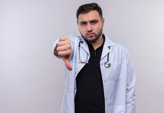 Jonge, bebaarde mannelijke arts die witte jas met stethoscoop draagt die ontevreden kijkt die afkeer toont