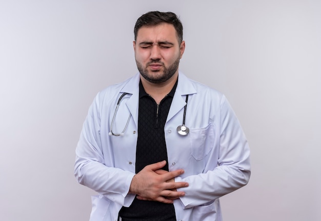 Jonge, bebaarde mannelijke arts die witte jas met stethoscoop draagt die onwel kijkt wat betreft zijn buik die pijn voelt