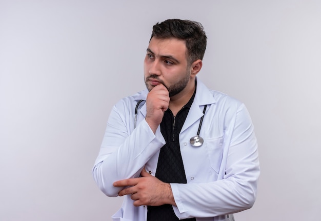 Jonge, bebaarde mannelijke arts die witte jas met stethoscoop draagt die opzij met hand op kin met peinzende uitdrukking op gezicht kijkt