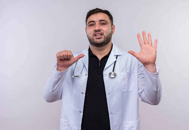 Jonge, bebaarde mannelijke arts die witte laag met stethoscoop draagt die en met vingers nummer zes benadrukt