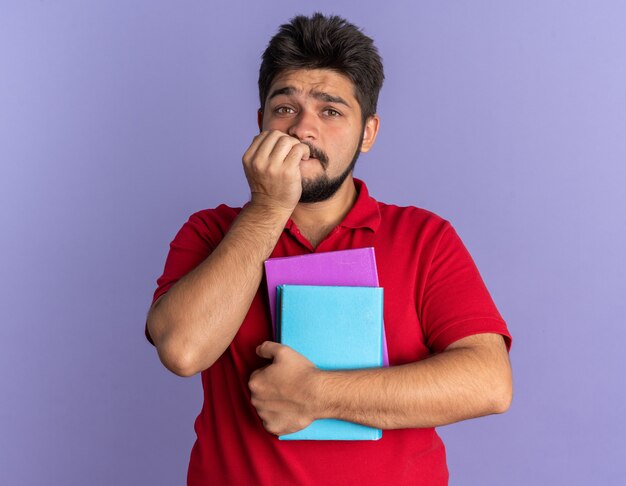 Jonge, bebaarde student in een rood poloshirt met boeken die gestrest en nerveus bijtende nagels over een blauwe muur staan