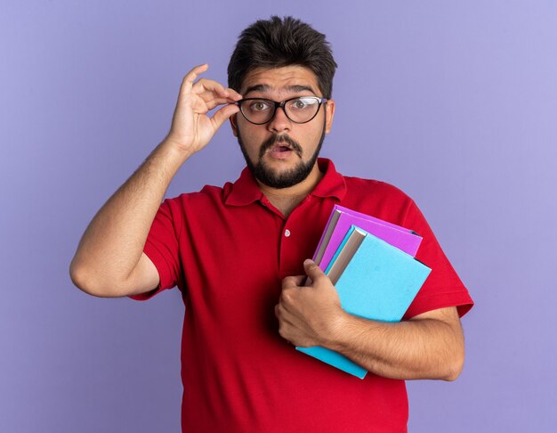 Jonge, bebaarde student in een rood poloshirt met een bril die boeken vasthoudt, verrast over de blauwe muur