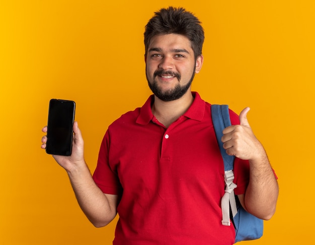 Jonge, bebaarde student in een rood poloshirt met rugzak die een smartphone vasthoudt en er glimlachend zelfverzekerd uitziet met zijn duimen omhoog staand
