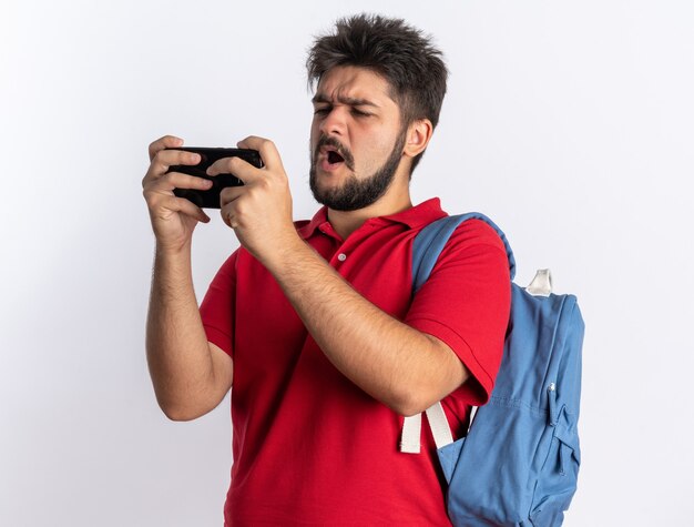 Jonge, bebaarde student in een rood poloshirt met rugzak die spelletjes speelt met een smartphone die er blij en opgewonden uitziet