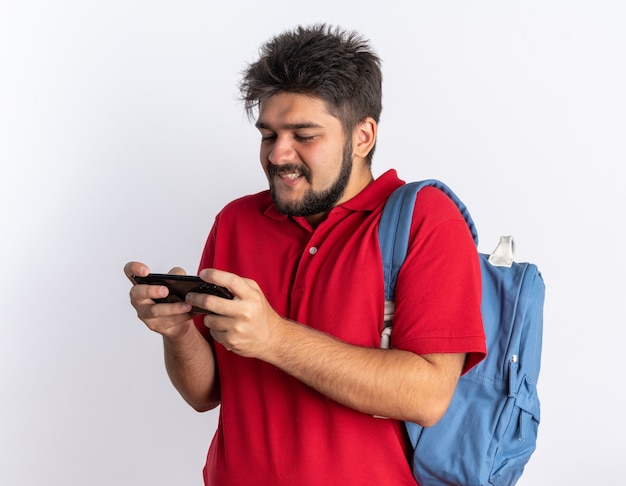 Jonge, bebaarde student in een rood poloshirt met rugzak die spelletjes speelt met smartphone gelukkig en vrolijk staand