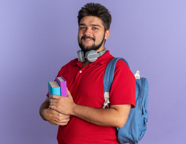 Jonge, bebaarde student in een rood poloshirt met rugzak met boeken met een koptelefoon om de nek en ziet er gelukkig en positief glimlachend uit