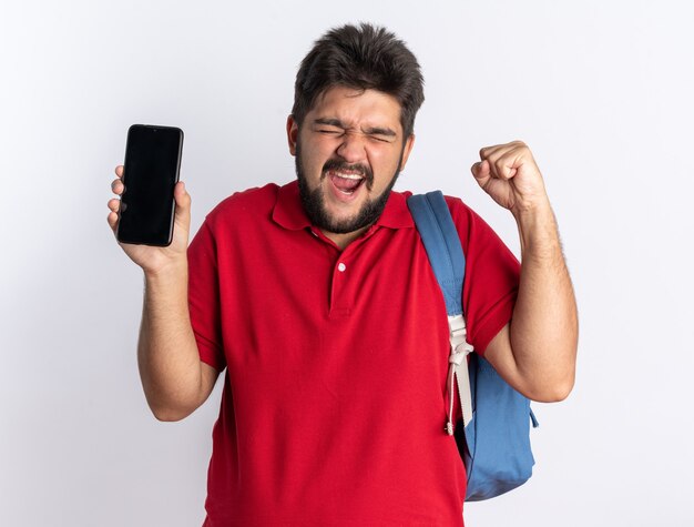 Jonge, bebaarde student in een rood poloshirt met rugzak met een smartphone die zijn vuist balde, blij en opgewonden staand