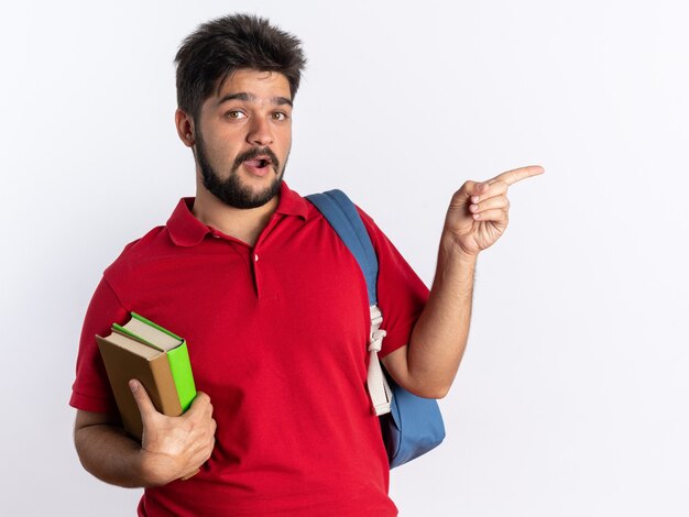 Jonge, bebaarde student in een rood poloshirt met rugzak met notebooks die er blij en verrast uitziet en met de wijsvinger naar de zijkant wijst