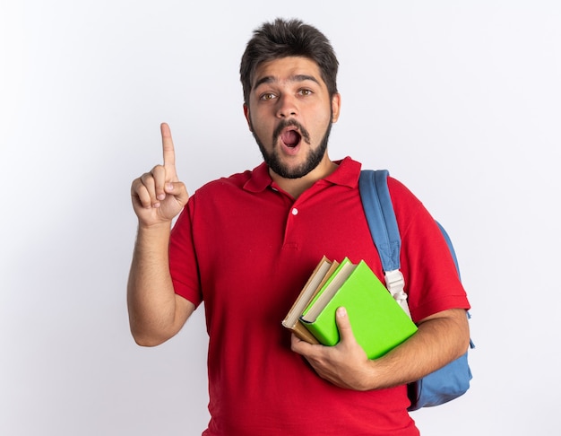 Jonge, bebaarde student in een rood poloshirt met rugzak met notitieboekjes die er blij en verrast uitziet met een wijsvinger nieuw idee concept staand
