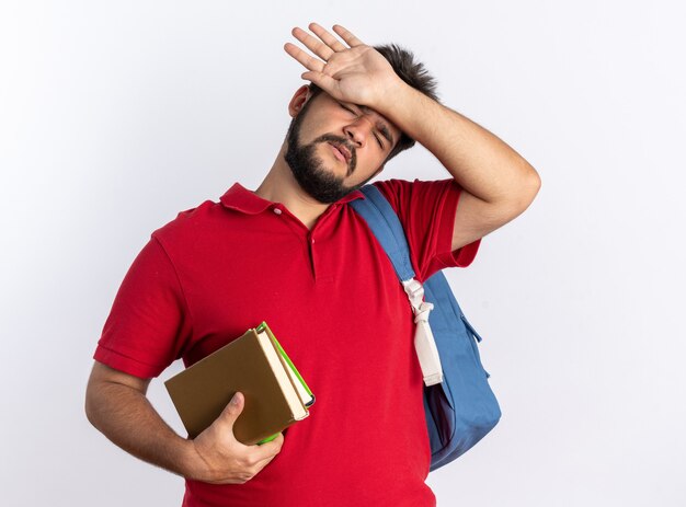 Jonge, bebaarde student in een rood poloshirt met rugzak met notitieboekjes die er moe en overwerkt uitziet en over een witte muur staat