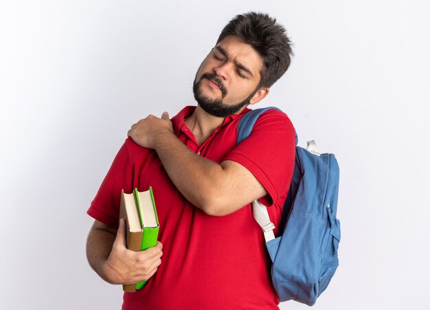 Jonge, bebaarde student in een rood poloshirt met rugzak met notitieboekjes die er niet goed uitziet en zijn schouder aanraakt, pijn voelend