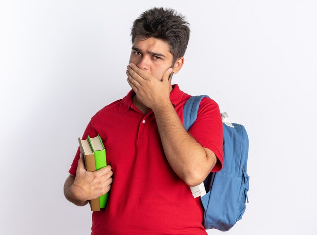 Jonge, bebaarde student in een rood poloshirt met rugzak met notitieboekjes die geschokt is en zijn mond bedekt met de hand die over een witte muur staat