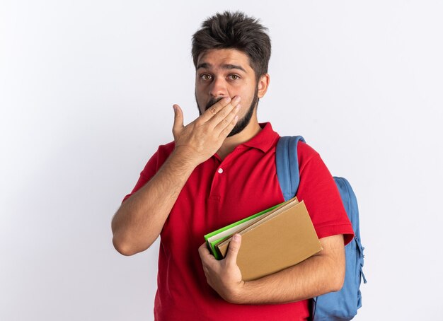 Jonge, bebaarde student in een rood poloshirt met rugzak met notitieboekjes die geschokt is en zijn mond bedekt met de hand die over een witte muur staat