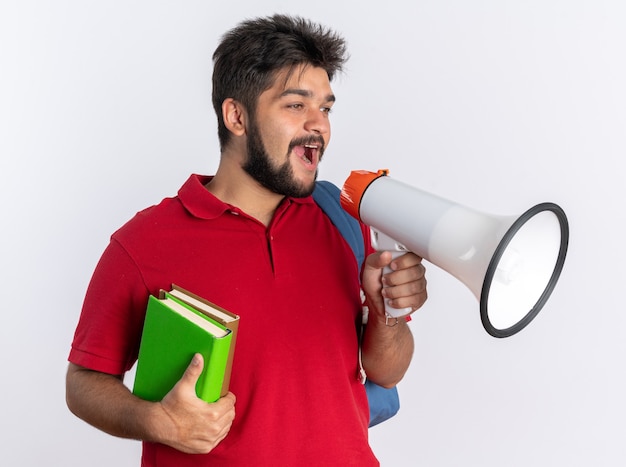 Jonge, bebaarde student in een rood poloshirt met rugzak met notitieboekjes die naar megafoon schreeuwt, blij en positief